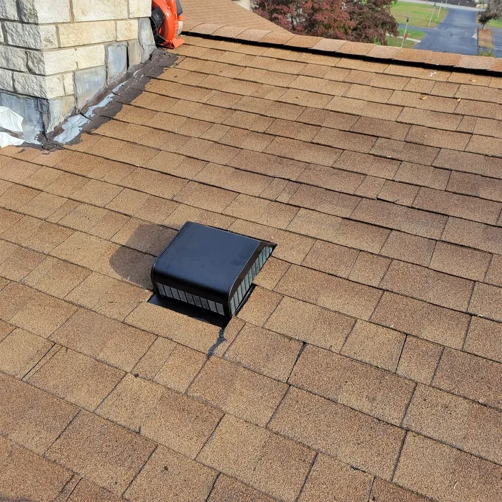 A black box roof vent installed on brown asphalt shingles near a stone chimney.