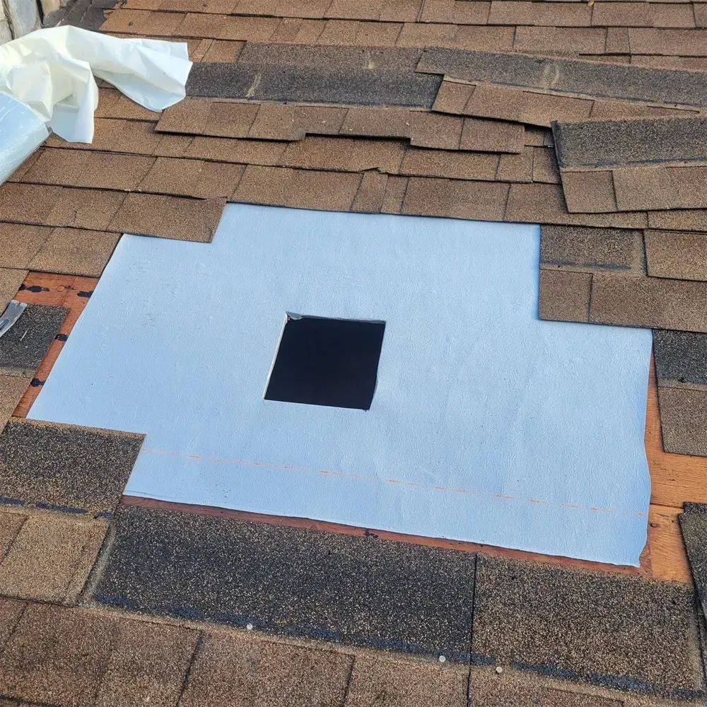 A patch of light gray underlayment on a roof with a square hole cut in the center, surrounded by brown asphalt shingles.