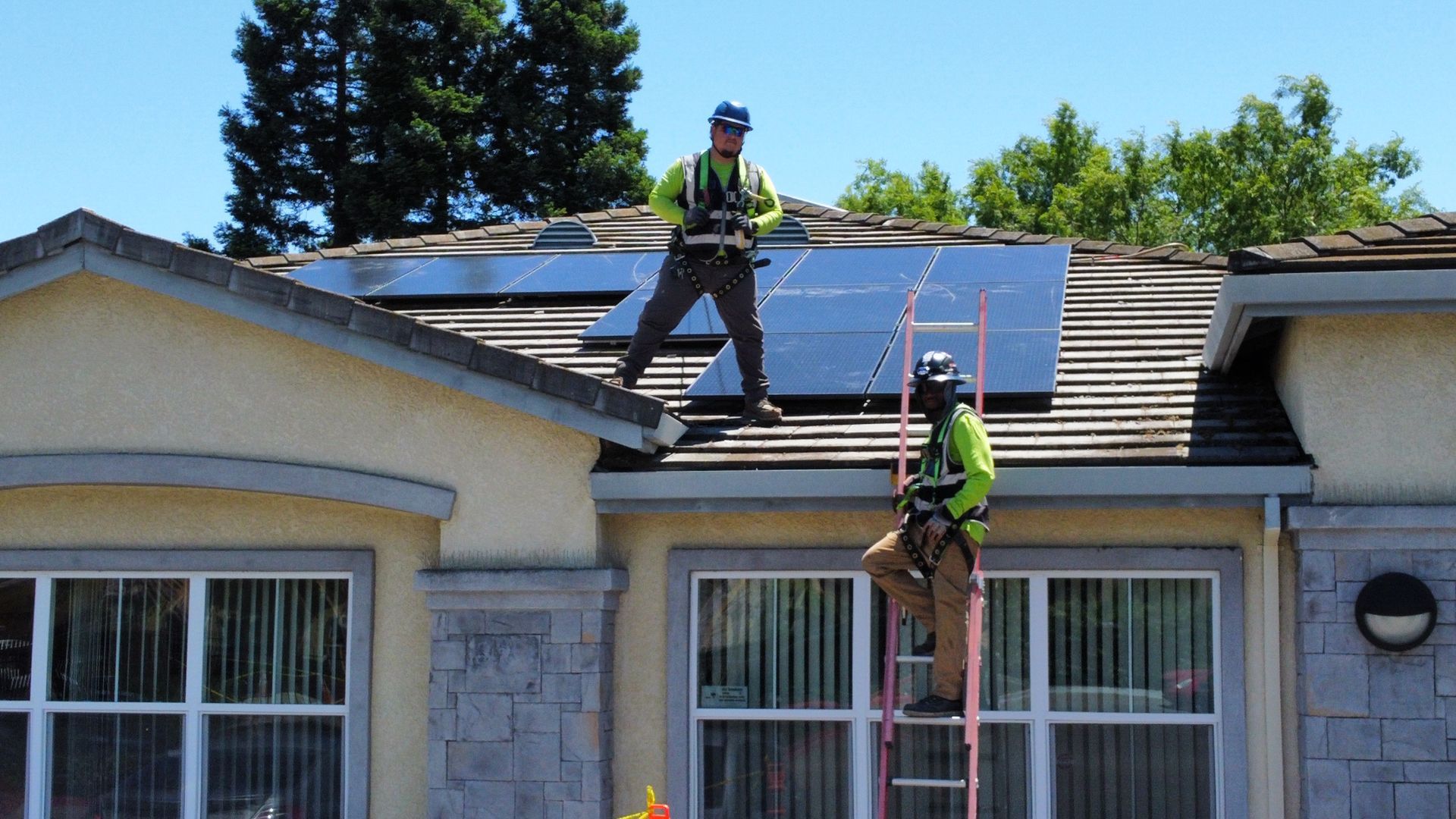 Roofers working on a residential roof, with a ladder and tools. Blue sky and foliage in the background.