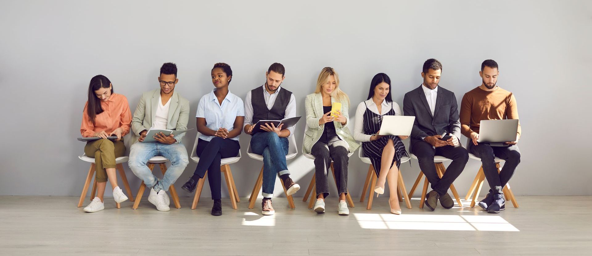 People sitting in a row, using laptops and tablets, waiting in a brightly lit room with a gray wall.