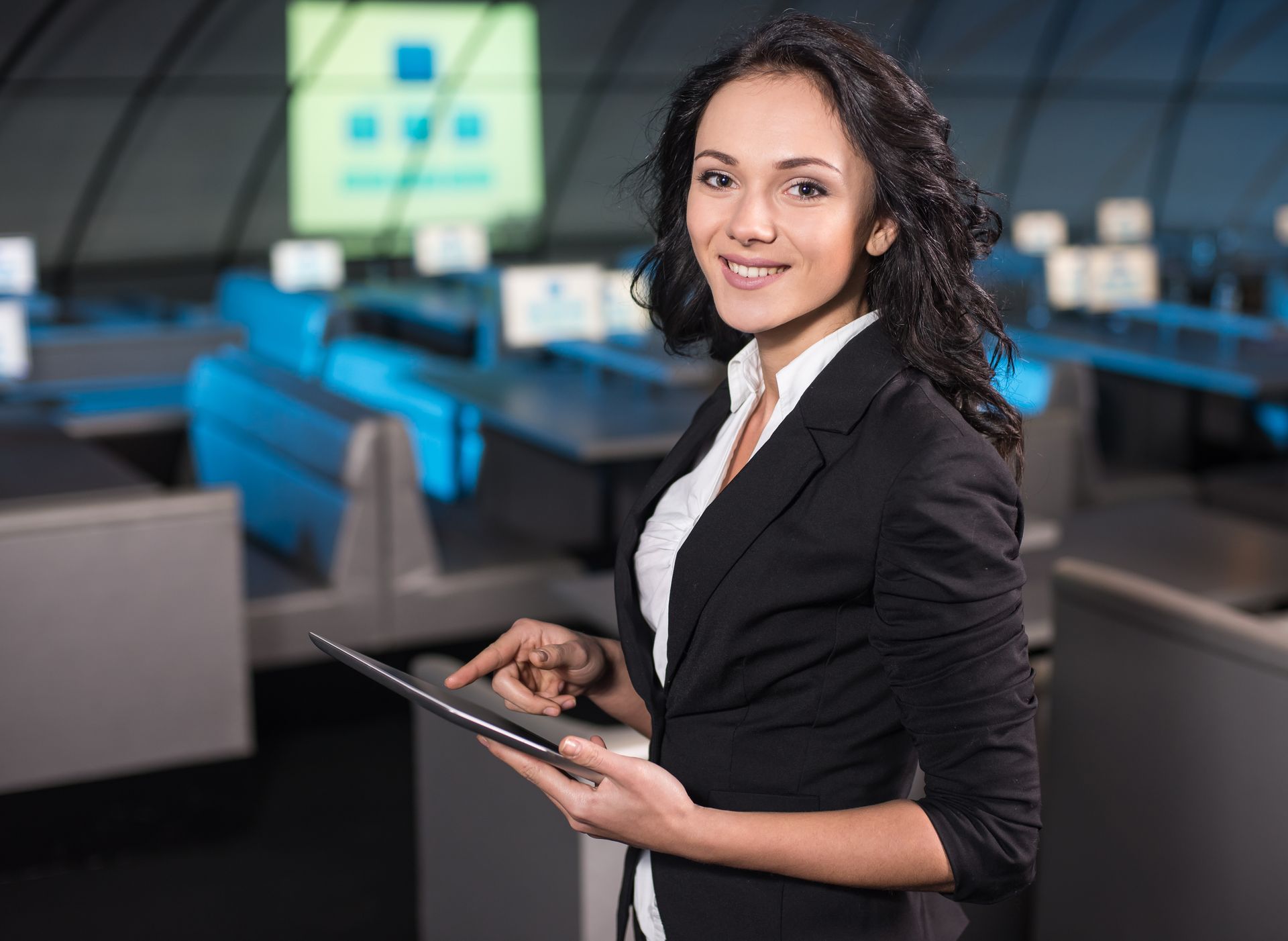 Woman in blazer holds tablet, smiling, in a technology-filled office setting.