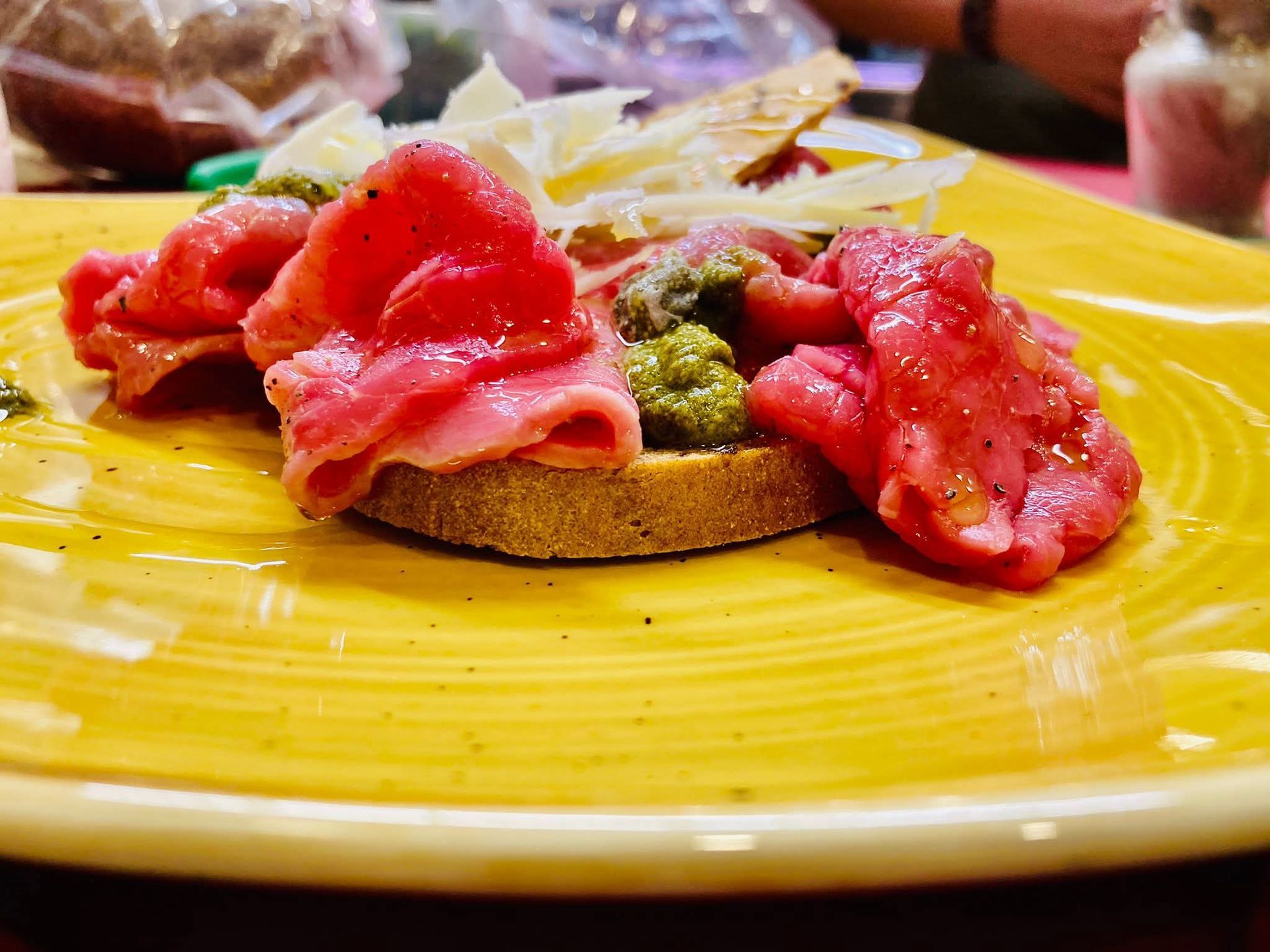 Carpaccio di manzo su pane tostato con pesto e parmigiano grattugiato, servito su un piatto giallo.