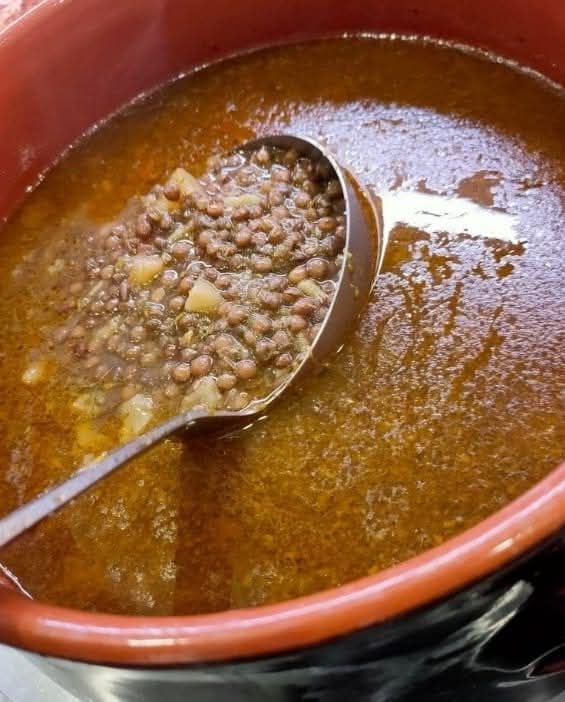 Zuppa di lenticchie in una ciotola di ceramica con un mestolo che raccoglie una porzione. Liquido marrone e lenticchie, ciotola arancione.