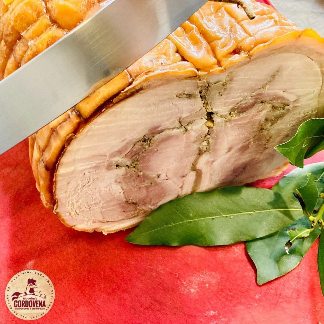 Un lombo di maiale arrosto tagliato a fette con un coltello, adagiato su una superficie rossa con foglie di alloro.