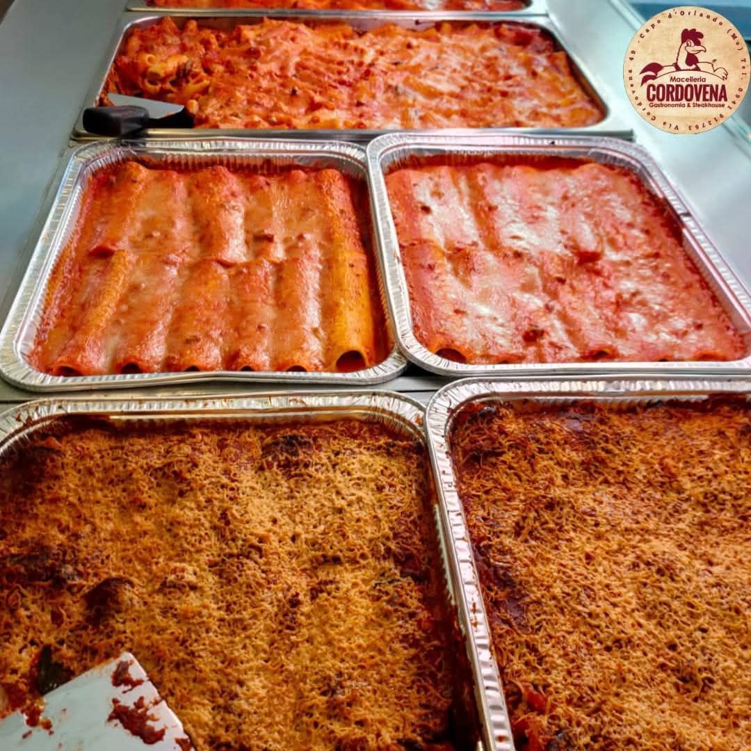 Vassoi di cibo italiano pronto: lasagne, cannelloni e pasta al forno, in un ambiente di cucina commerciale.