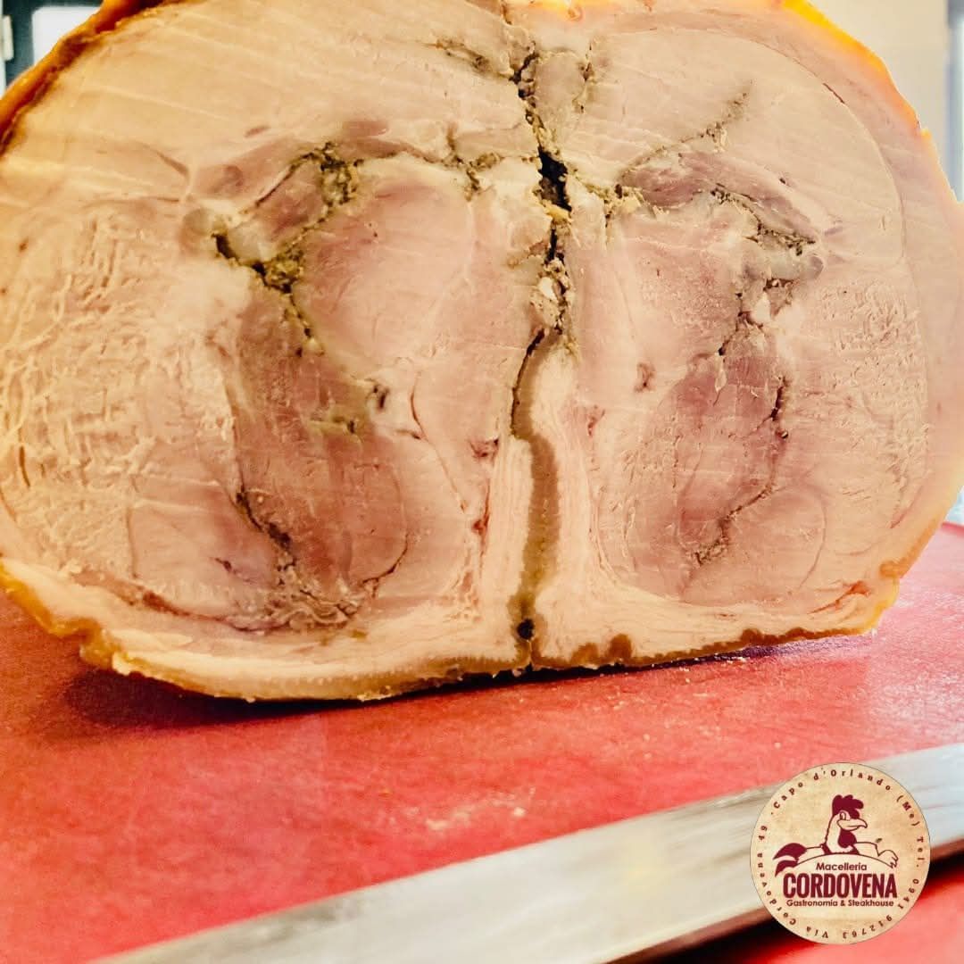 Sezione trasversale di maiale arrosto con erbe aromatiche visibili e cotenna croccante, su un tagliere rosso.