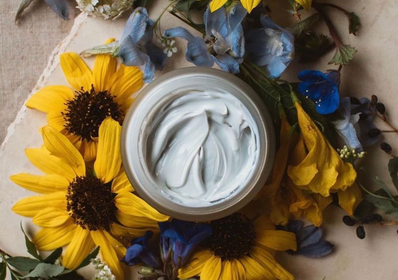 A jar of cream is surrounded by sunflowers and flowers.