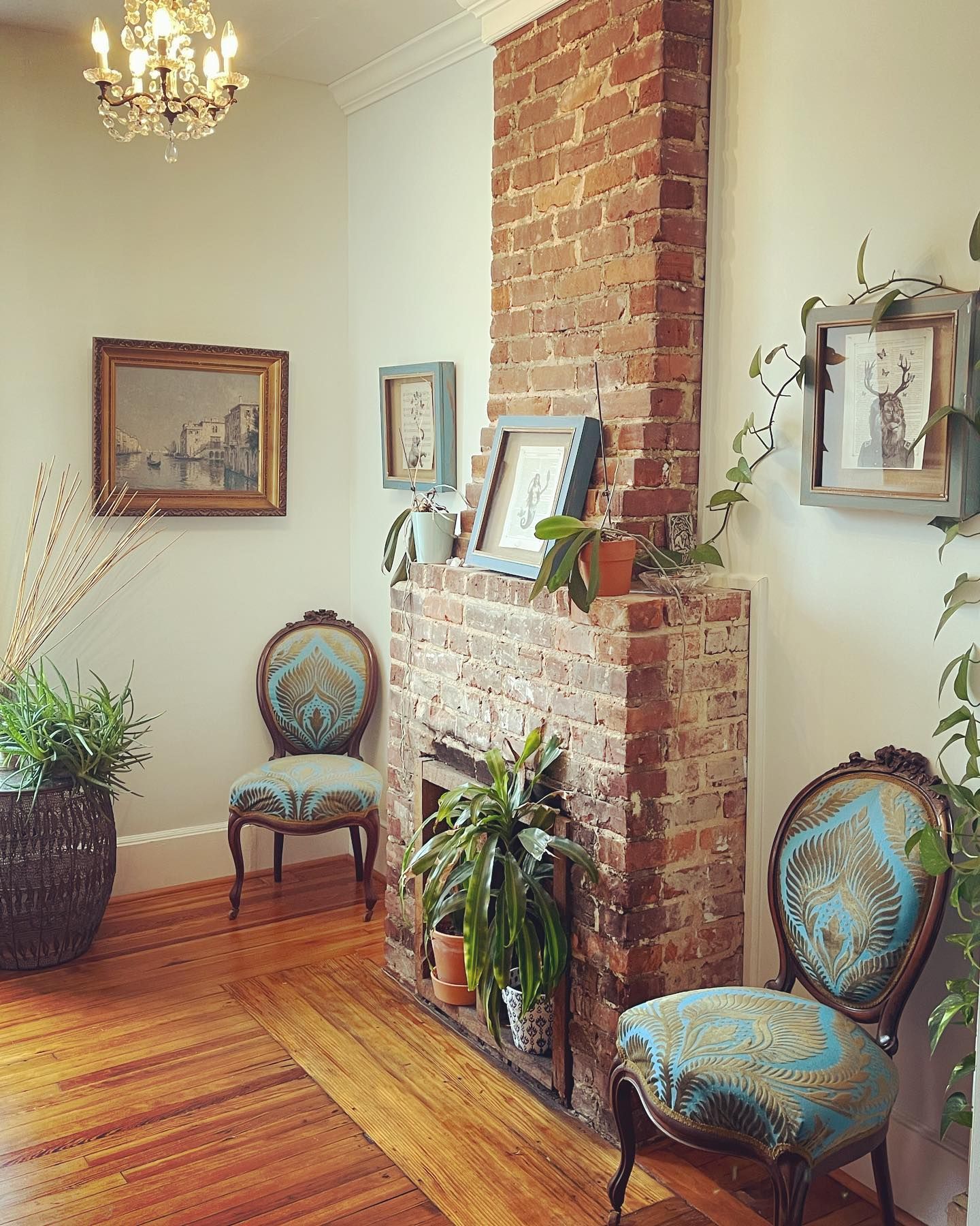 A living room with a brick fireplace , chairs , plants and a chandelier.