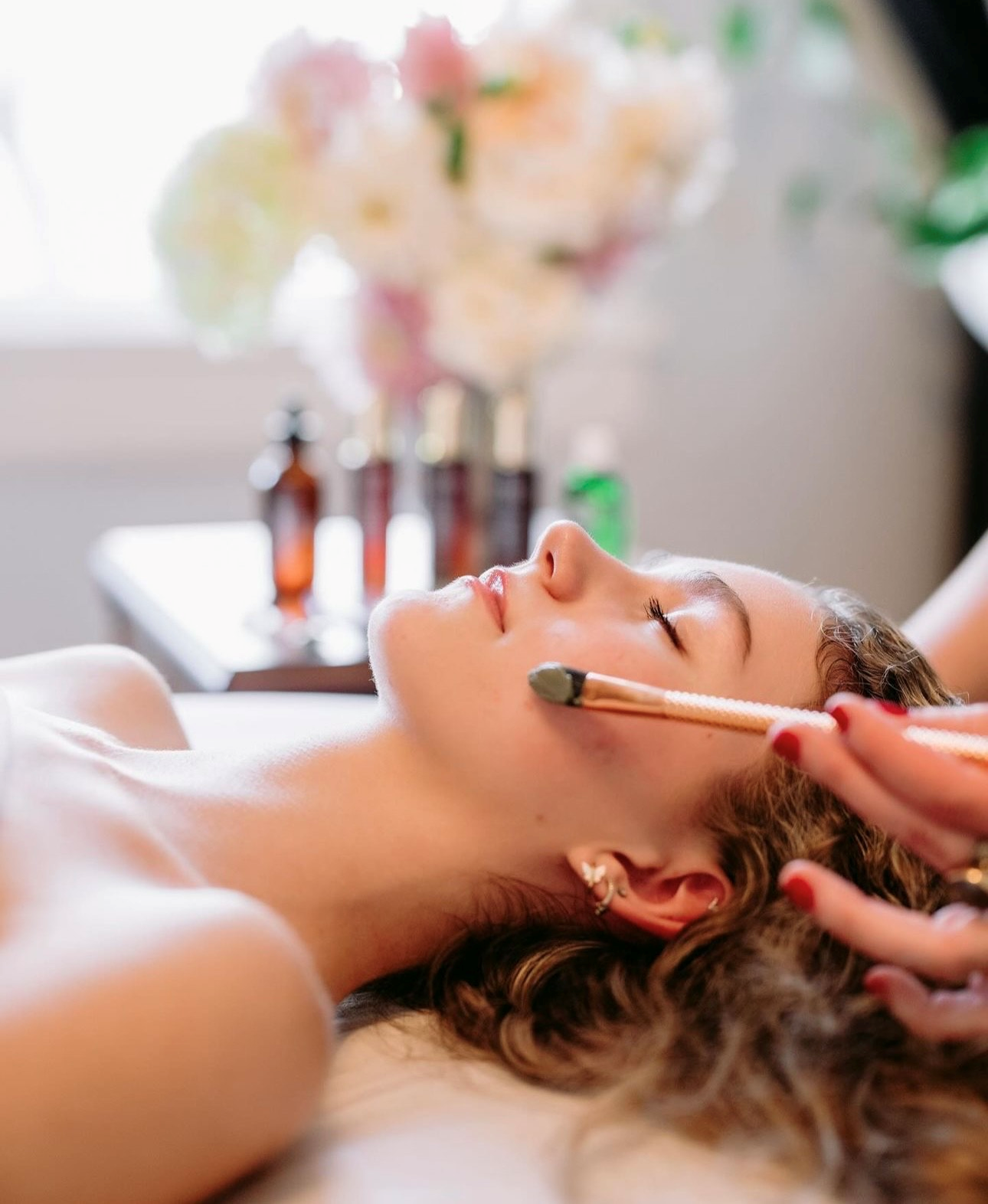 A woman is laying on a bed getting a facial treatment with a brush.