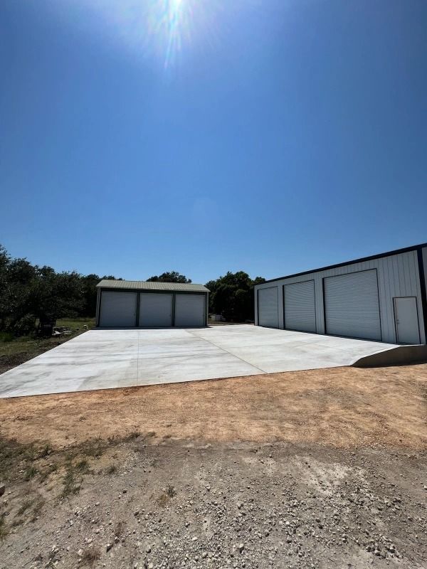 Two metal storage buildings with white garage doors on concrete pads under a bright blue sky.