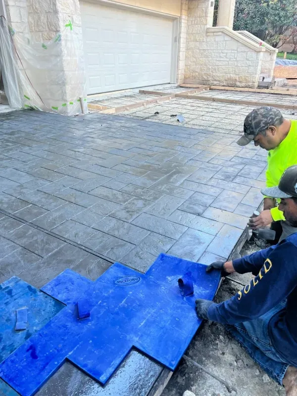 Two workers stamping concrete driveway with blue patterned forms near a garage.