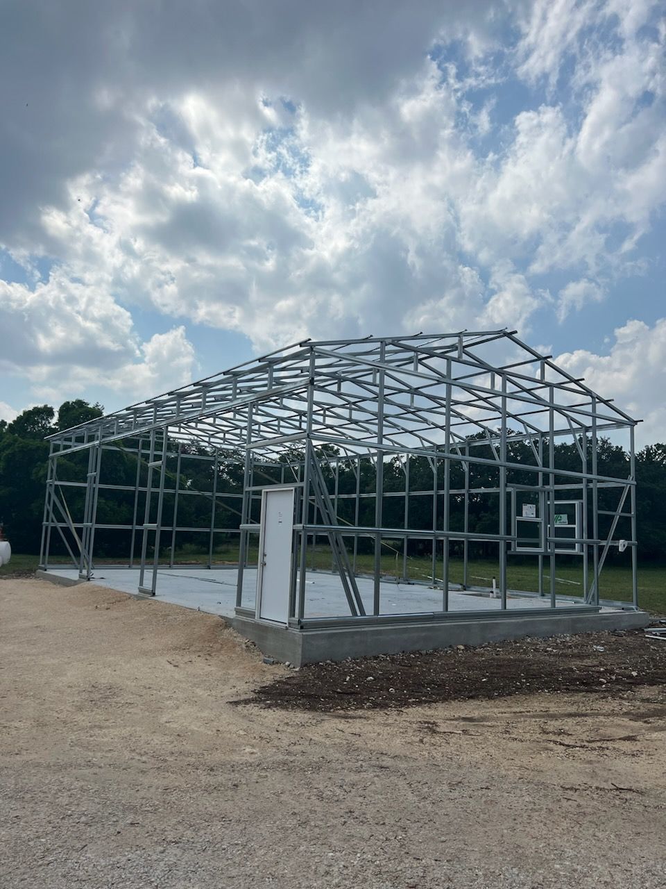 Metal frame structure of a building under construction, set on a concrete foundation, against a cloudy sky.
