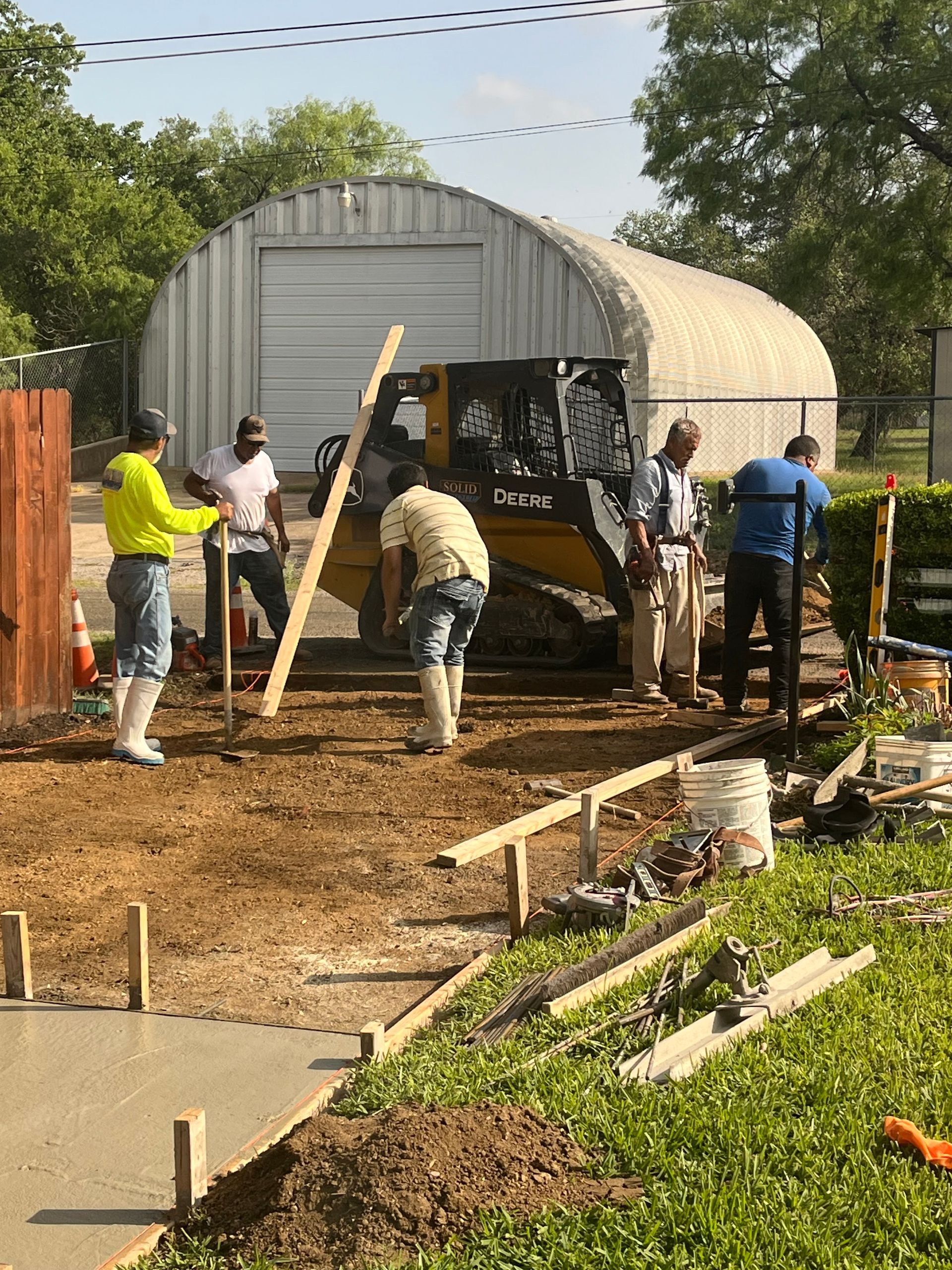 Construction workers paving a pathway with a skid steer loader near a metal building.