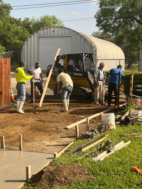 Construction workers paving a pathway with a skid steer loader near a metal building.
