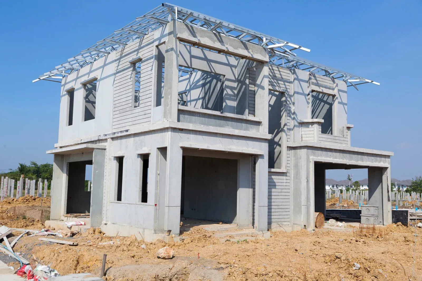 Two-story concrete house under construction; steel roof frame; dirt lot; blue sky.