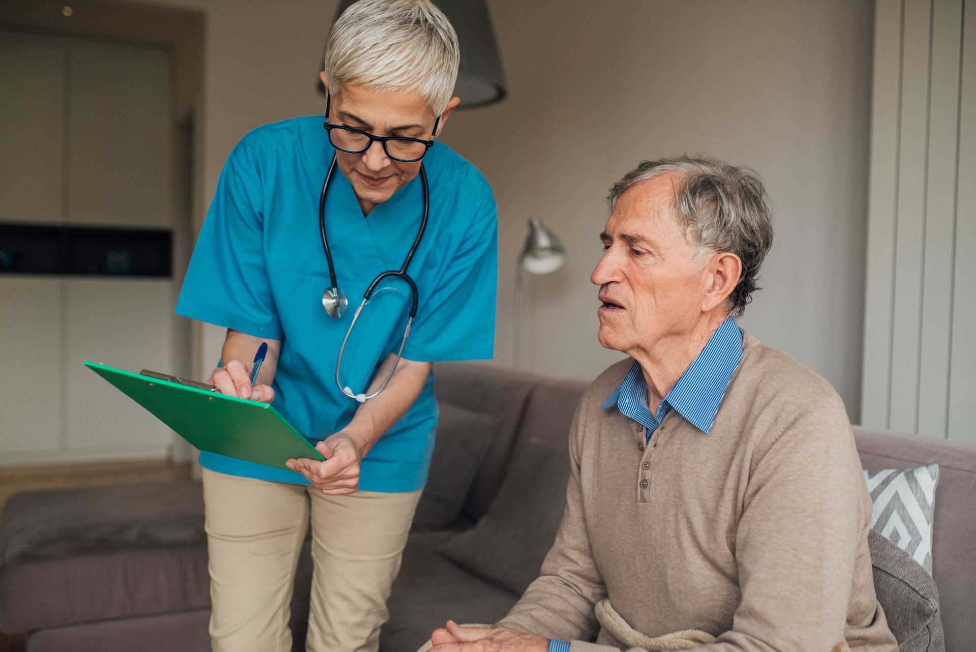Nurse in blue scrubs with stethoscope, writing on a clipboard while talking to an elderly person in a beige sweater seated on a couch.