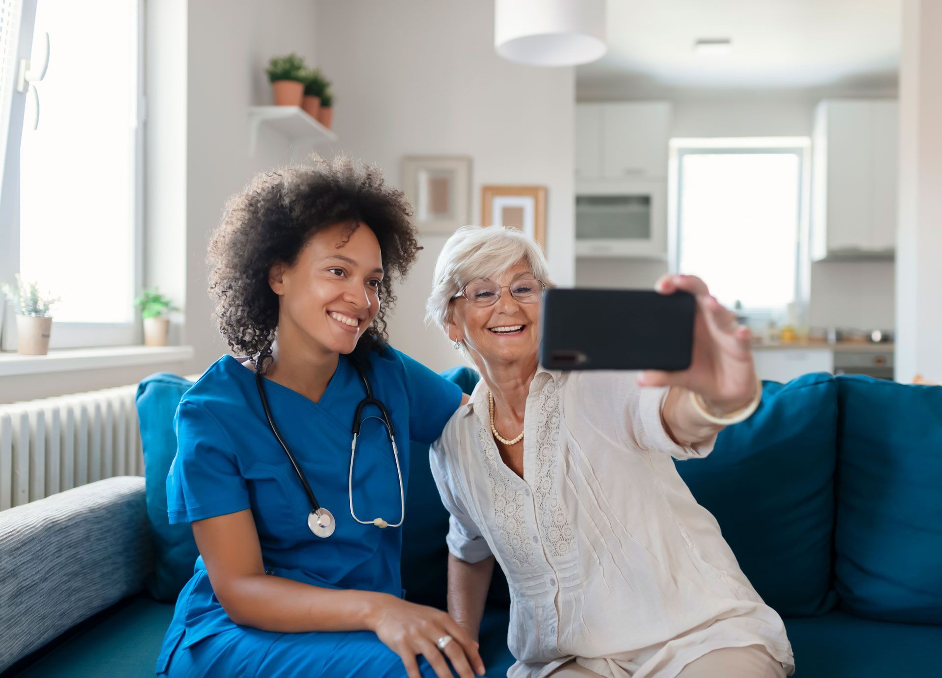 Caregiver in blue scrubs and senior woman taking selfie on a blue couch in a living room.