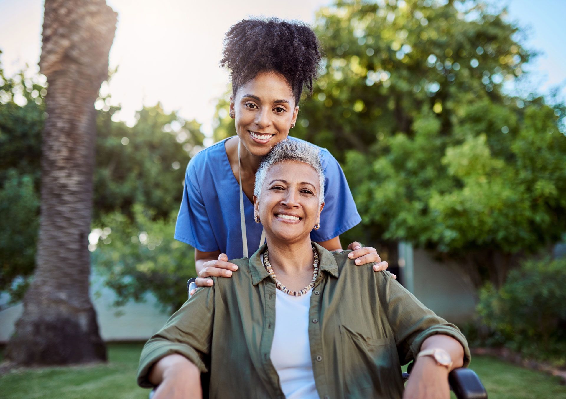 A young woman in blue scrubs assists an older woman in a wheelchair outdoors; both smile.