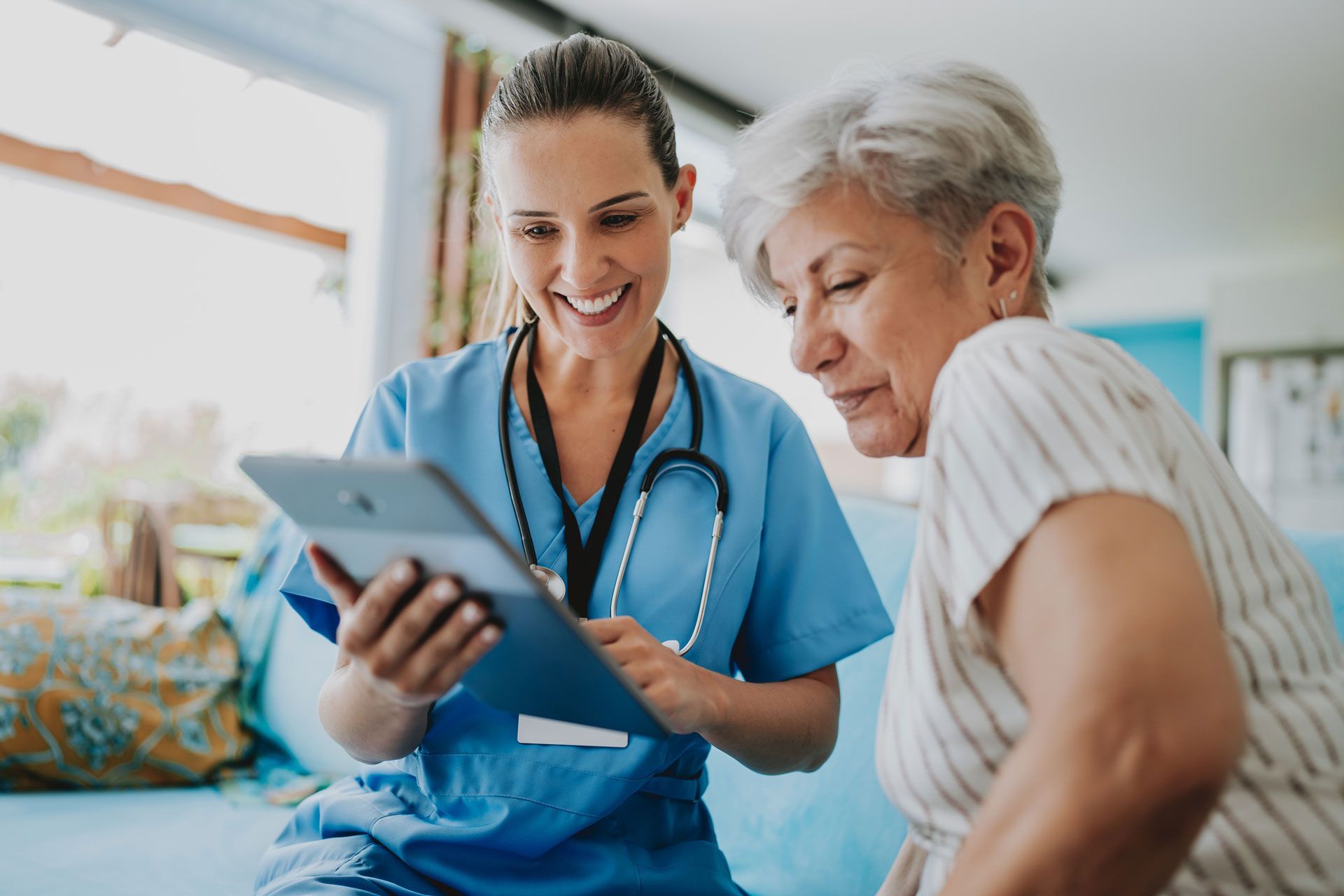 Nurse in blue scrubs shows a tablet to an older woman. Both smile, indoors with window.