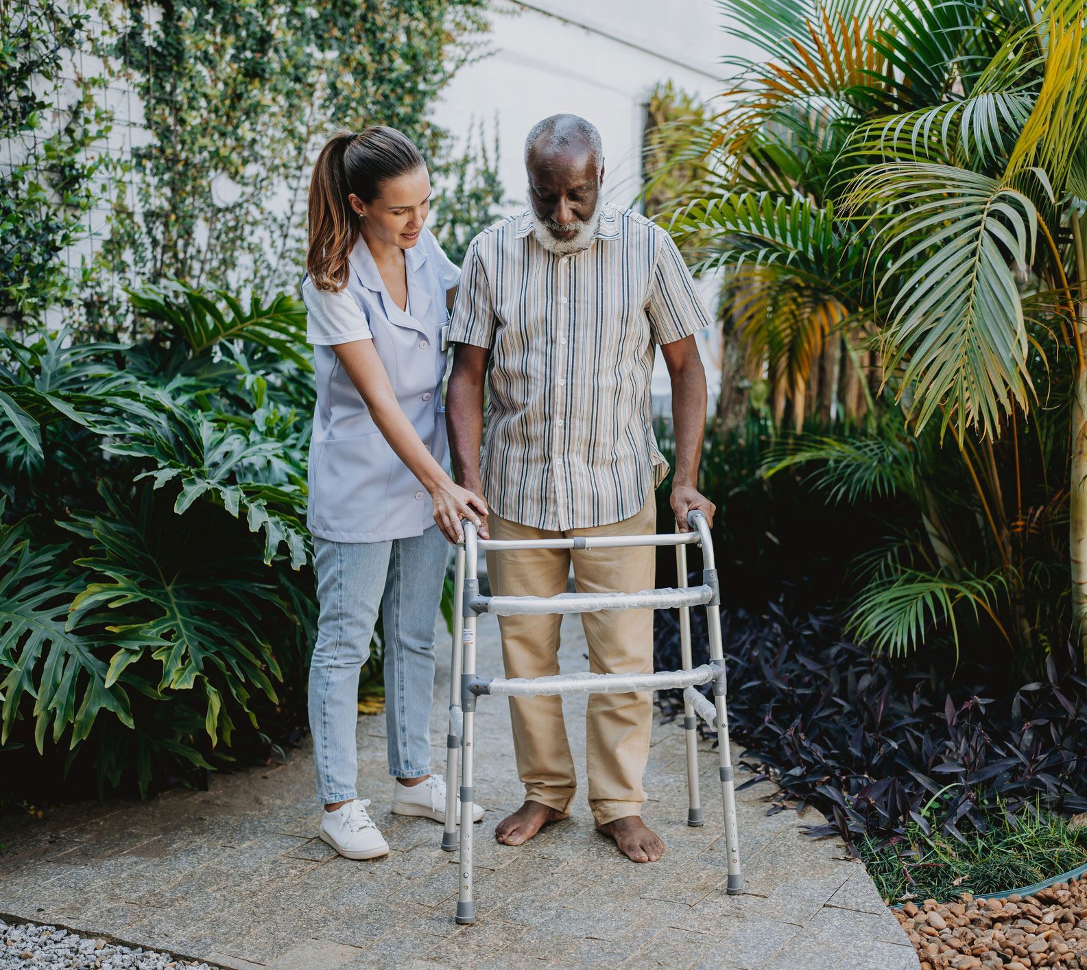 Caregiver assists senior using a walker on a stone path in a garden.