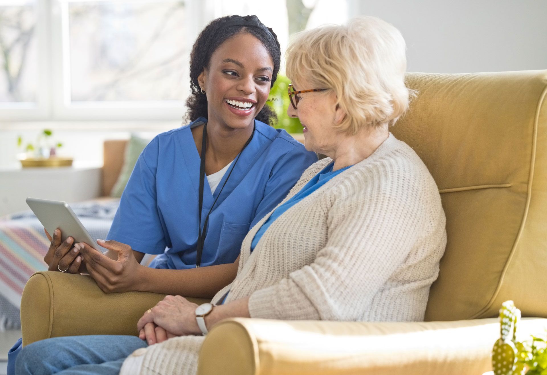 Nurse in blue scrubs smiles at elderly woman seated in a yellow chair, holding a tablet indoors.