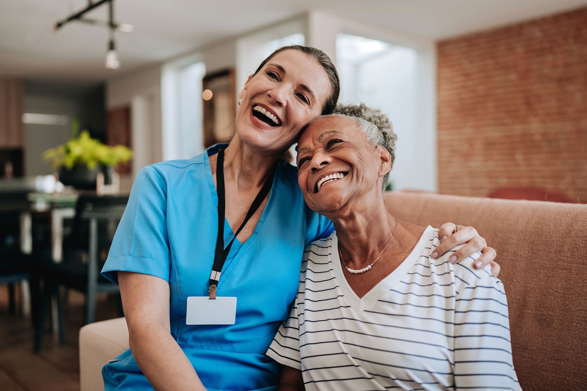 Woman in scrubs with her arm around smiling older woman on a couch.
