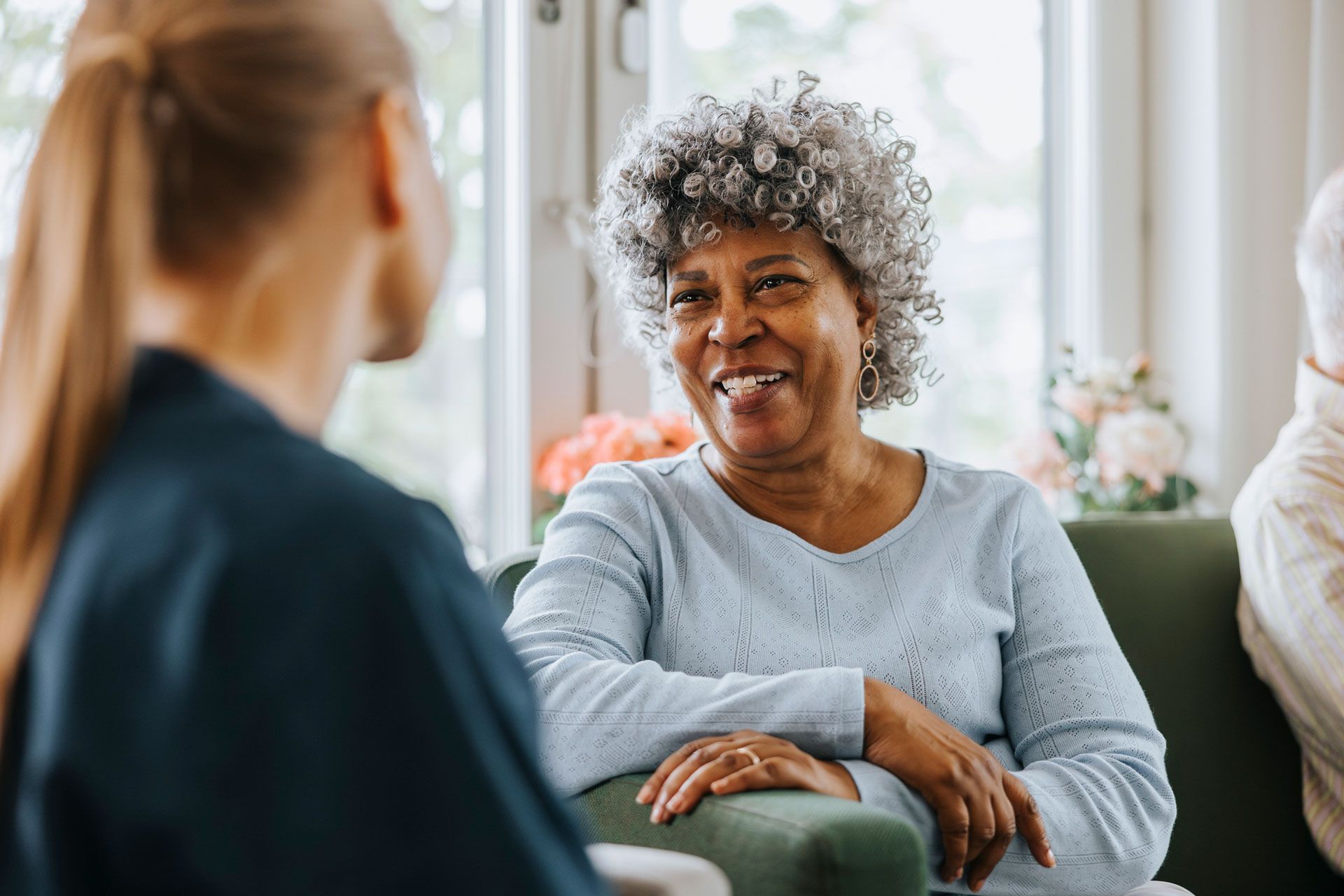Woman with gray hair smiles while talking to someone off-screen, sitting on a green couch.