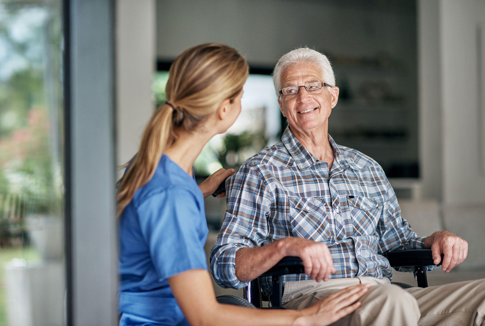 Nurse in blue scrubs assists a smiling elderly man in a wheelchair. Bright indoor setting.