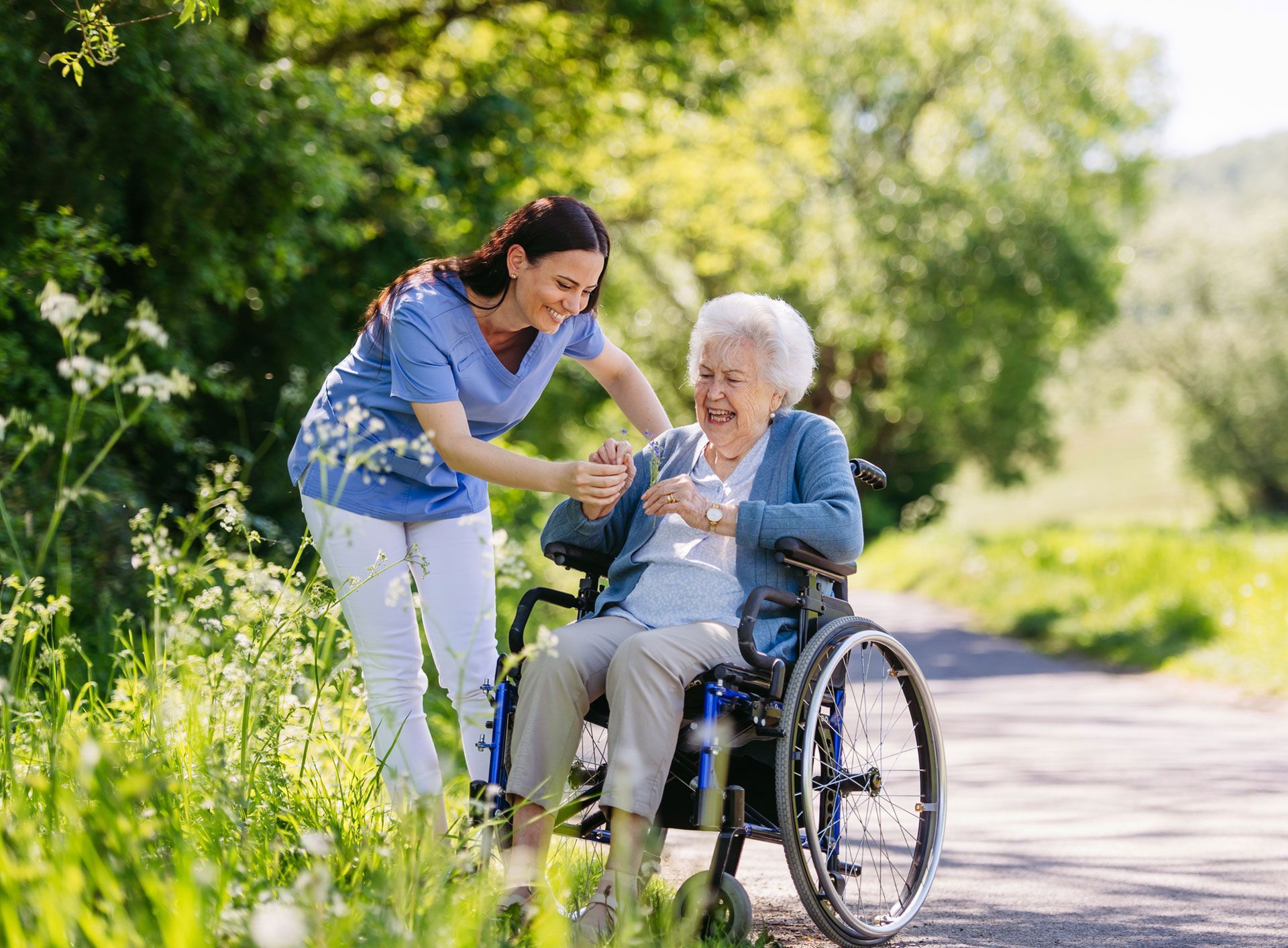 Caregiver helping an elderly woman in a wheelchair on a sunny outdoor path.