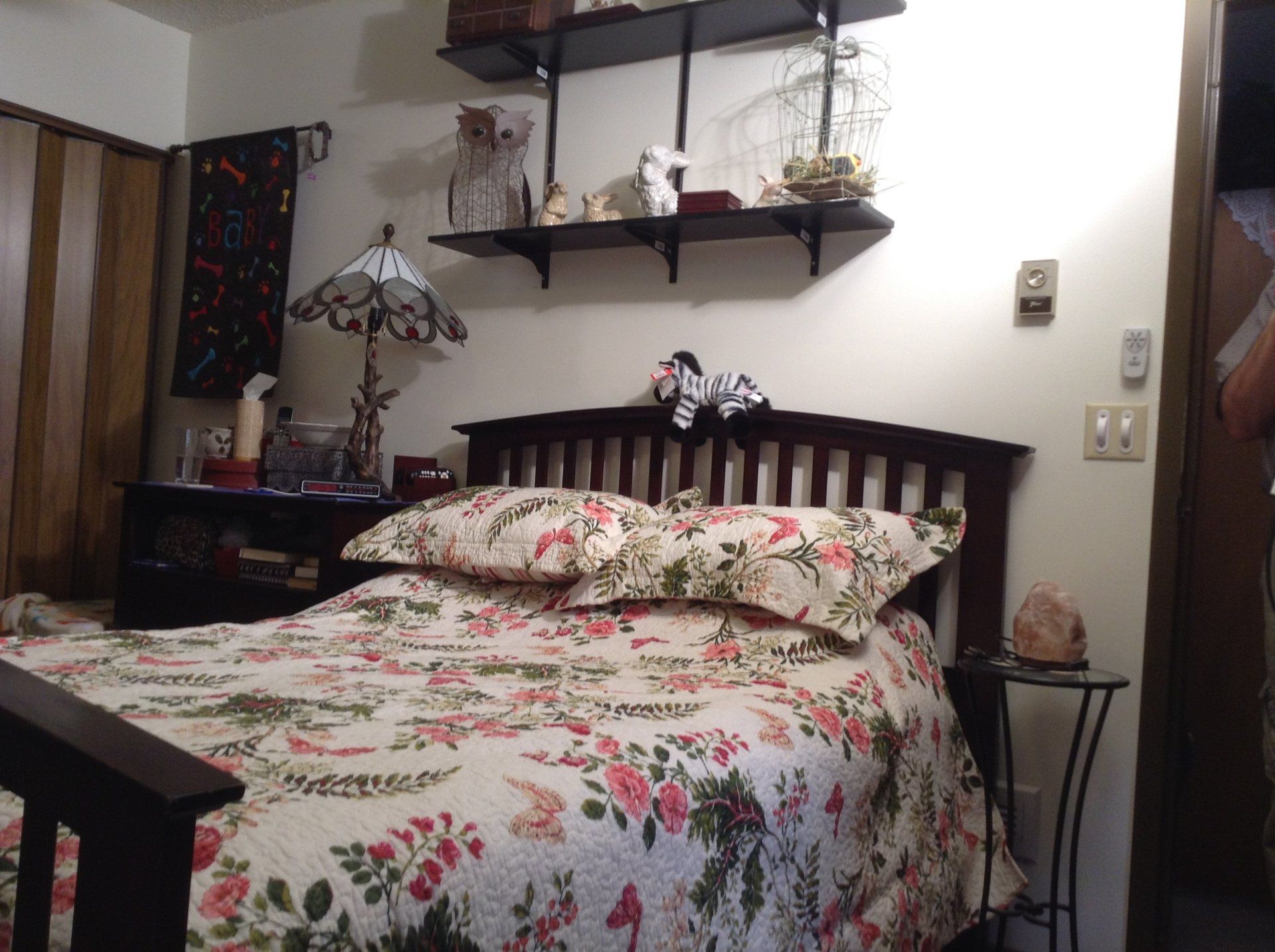 Bedroom with floral quilt, dark wooden bed frame, shelves, and lamp.