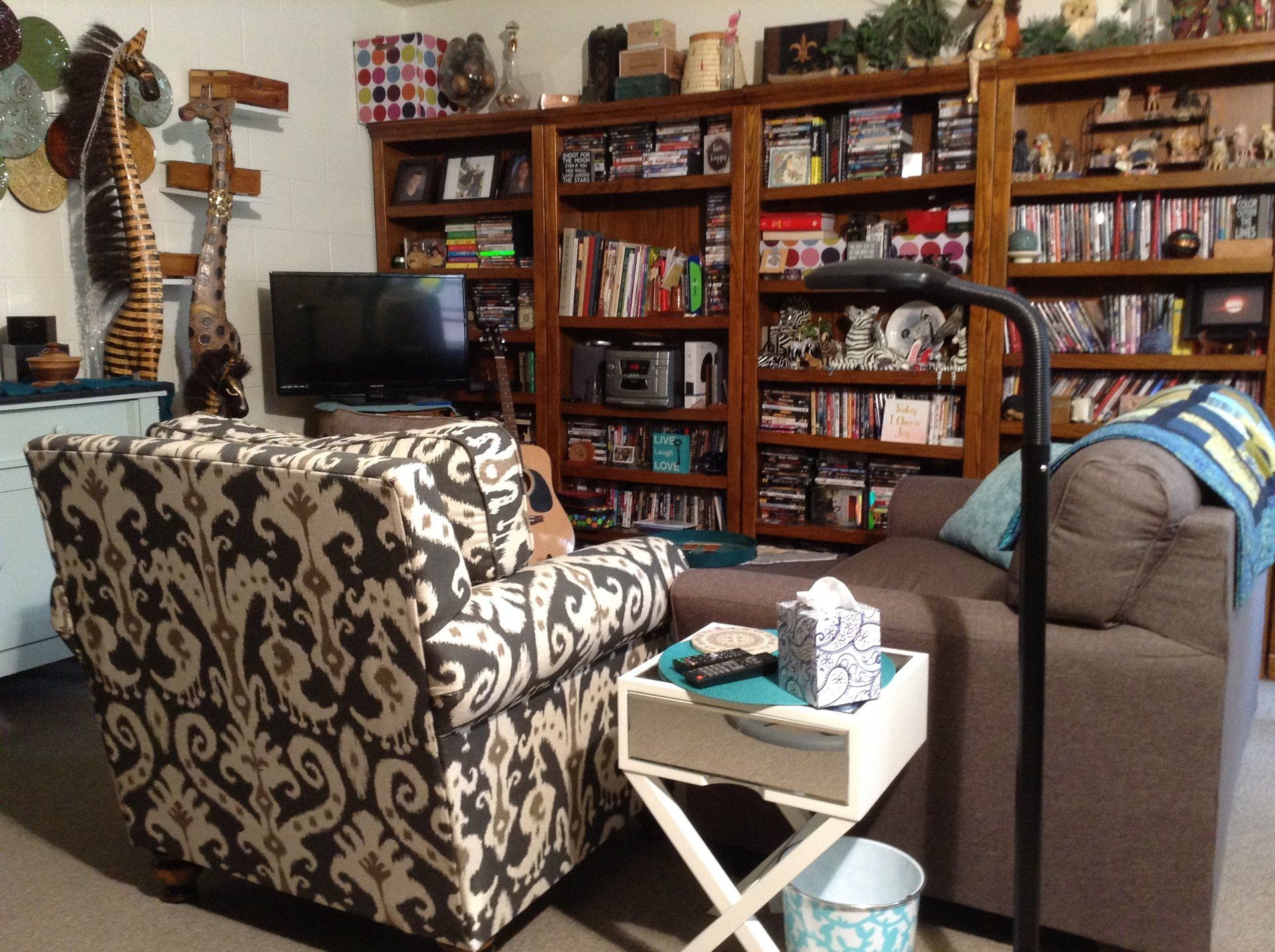 Cozy living room with patterned armchair, sofa, bookshelves filled with items, and a lamp.