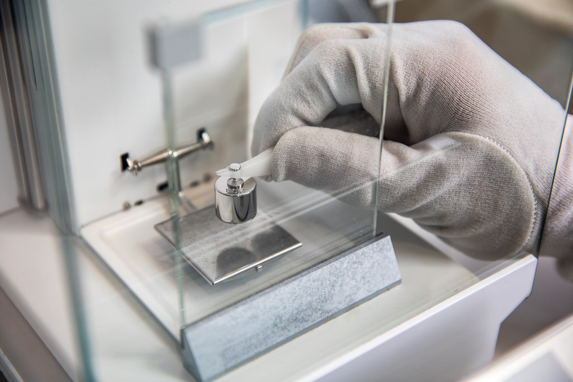 Gloved hand placing a weight on a precision balance scale in a laboratory setting.
