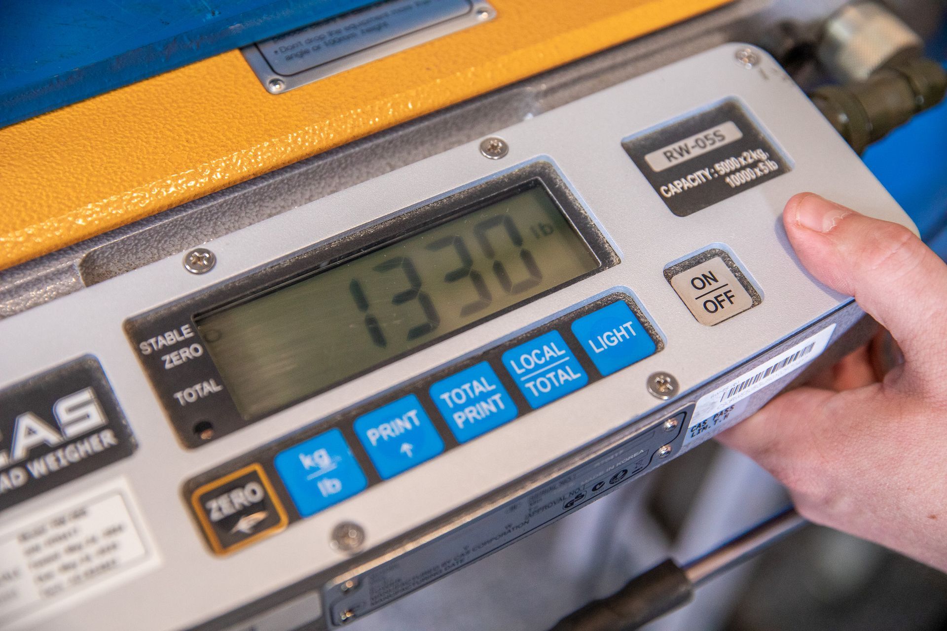 Hand holding a digital scale displaying 1330. The scale has buttons and a blue and yellow casing.