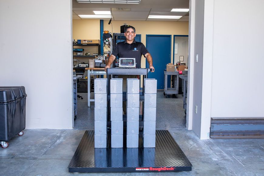 Man standing on a scale, smiling. Grey concrete blocks are stacked on the scale in a workshop.