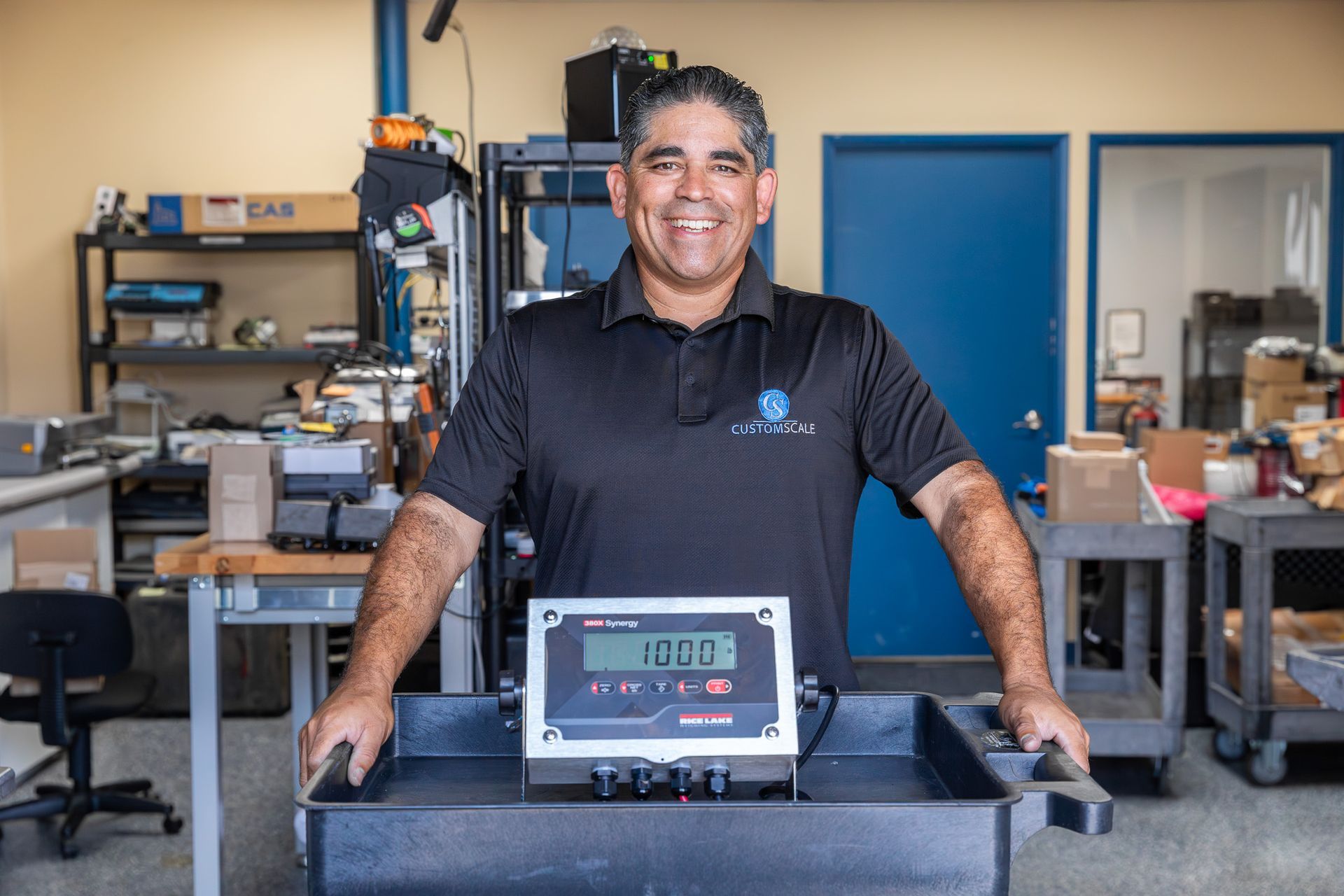 Man smiling, holding a weighing scale in a workshop.