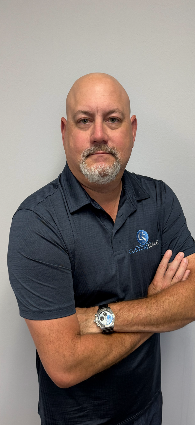 A bald man with a graying beard, wearing a dark blue polo shirt, arms crossed, and a watch.