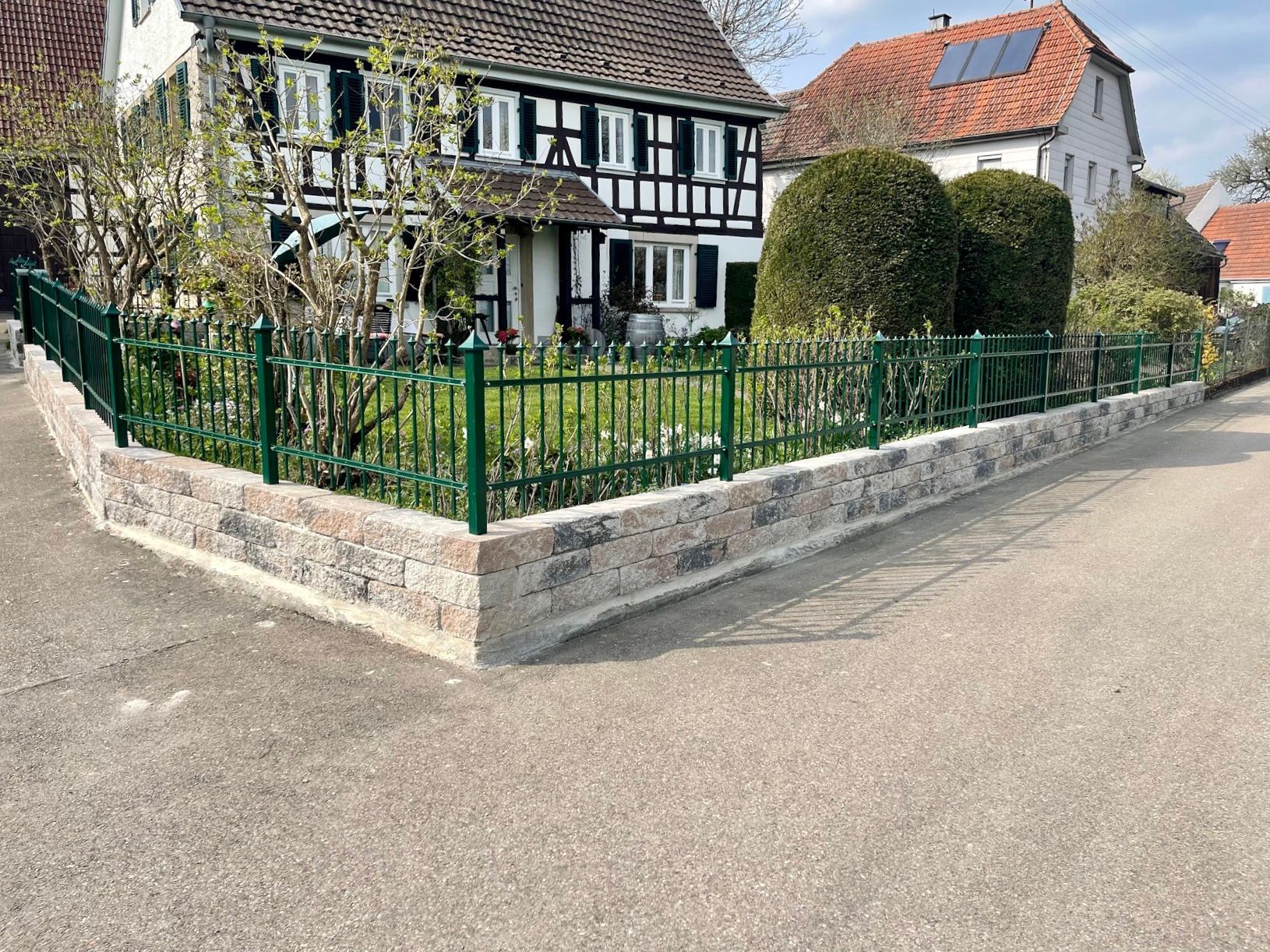 Ein grüner Zaun umgibt eine Steinmauer vor einem Haus.