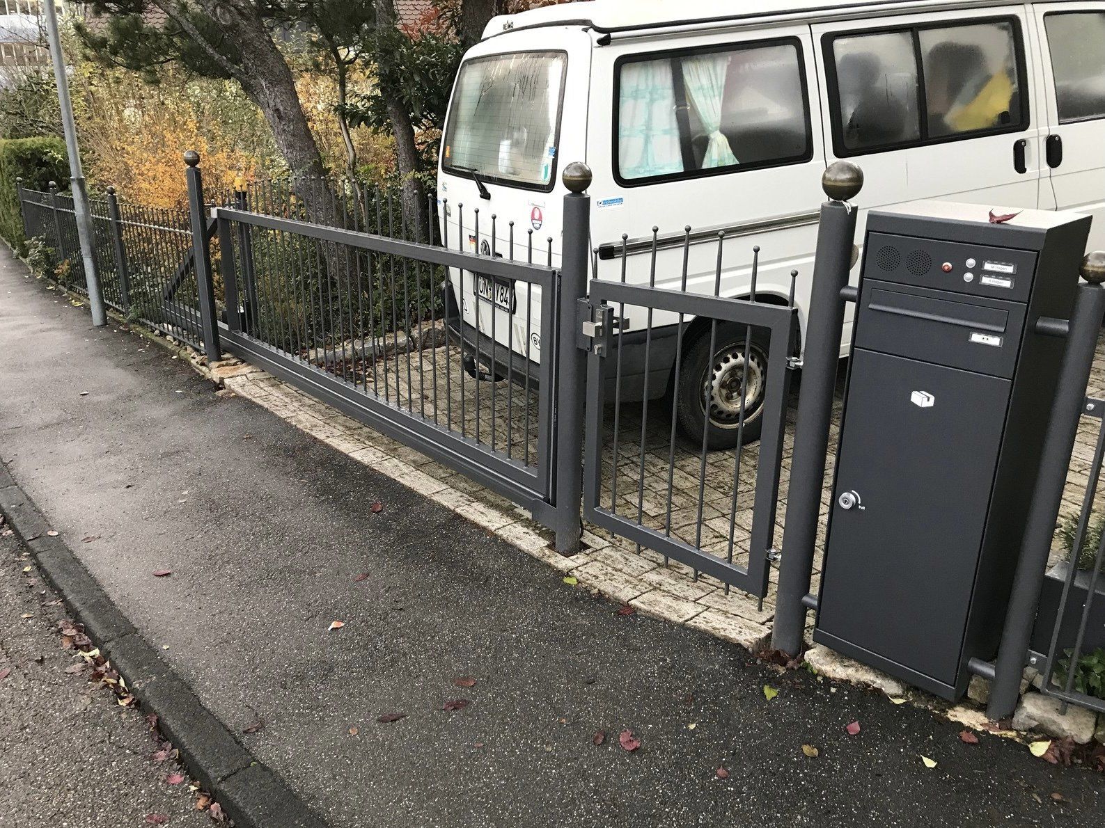 Ein weißer Lieferwagen steht hinter einem Zaun neben einem Briefkasten.
