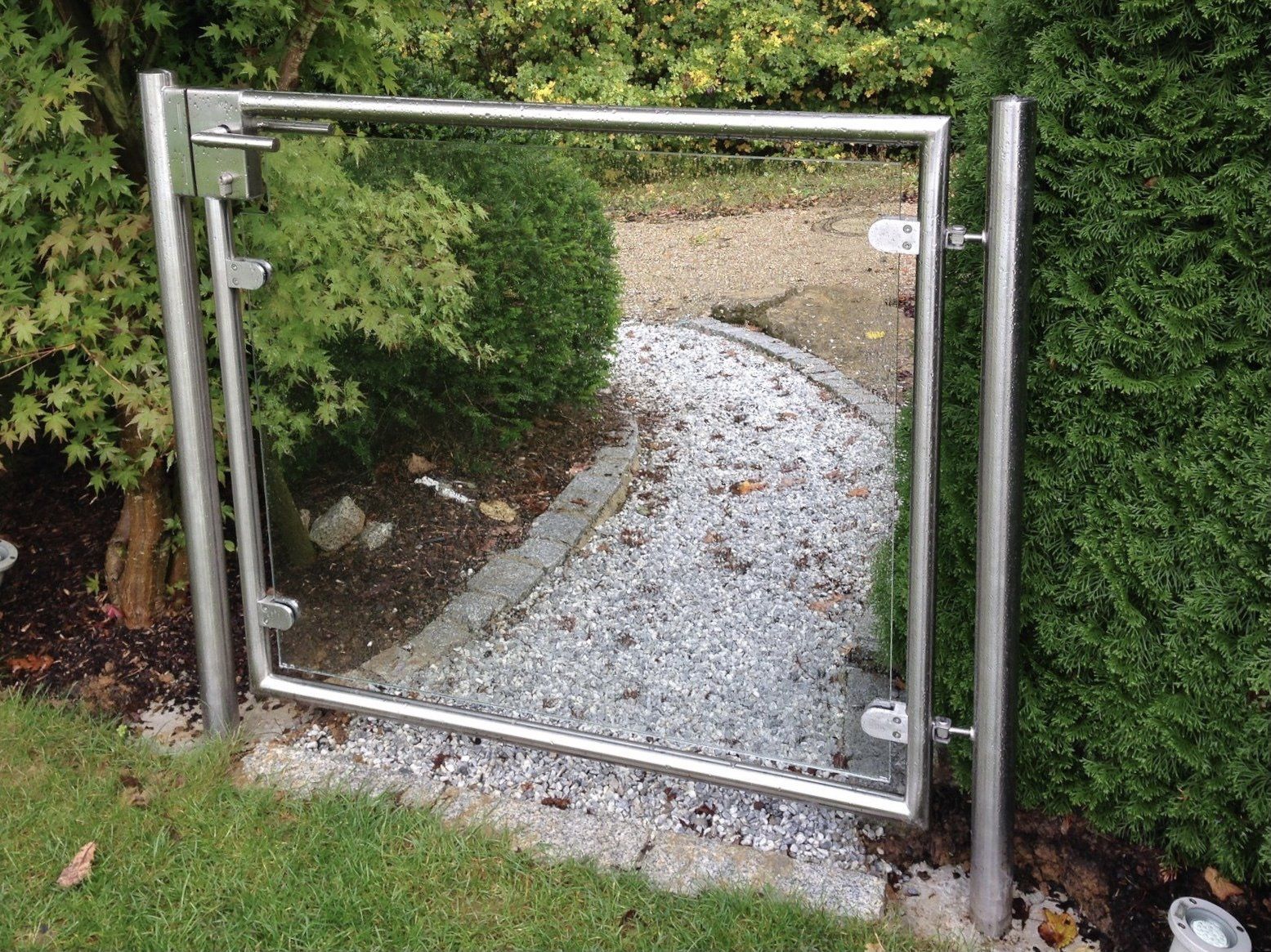 Ein Edelstahltor mit einer Glastür ist von Kies und Bäumen umgeben.