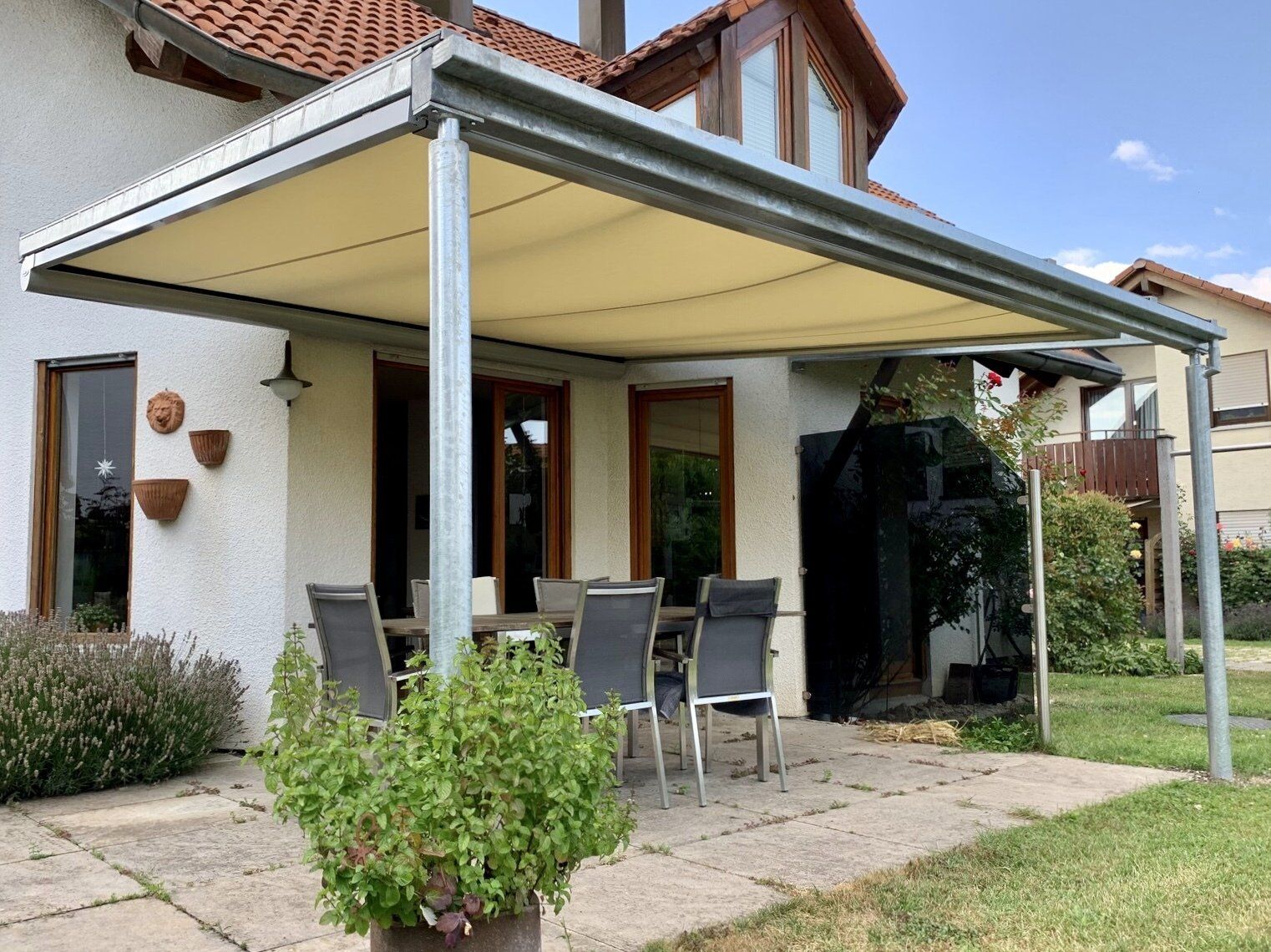 Eine Terrasse mit Tisch und Stühlen unter einem Vordach vor einem Haus.