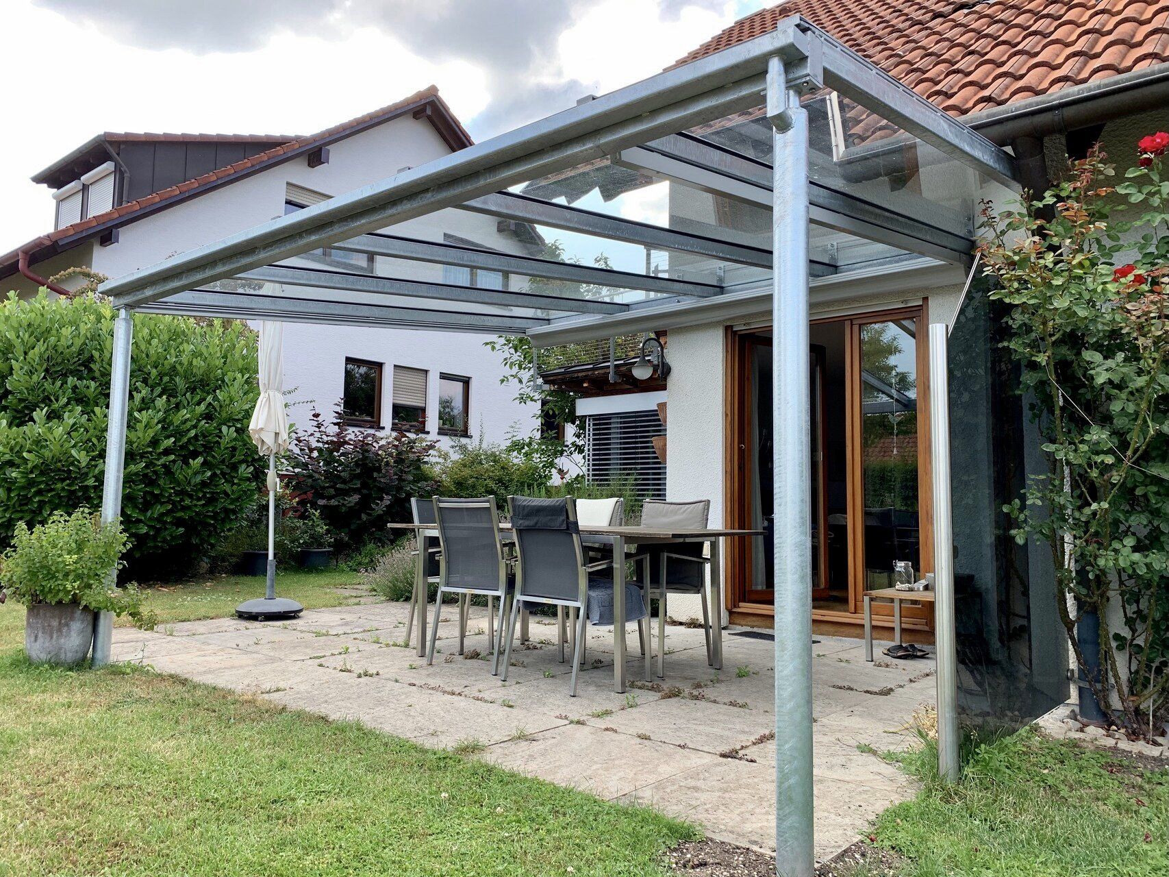Eine Terrasse mit Tisch und Stühlen unter einem Vordach vor einem Haus.