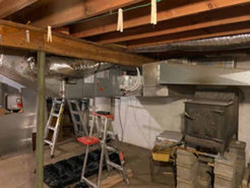 Basement interior with furnace, wood stove, and ductwork. Ladders and support beams are visible.