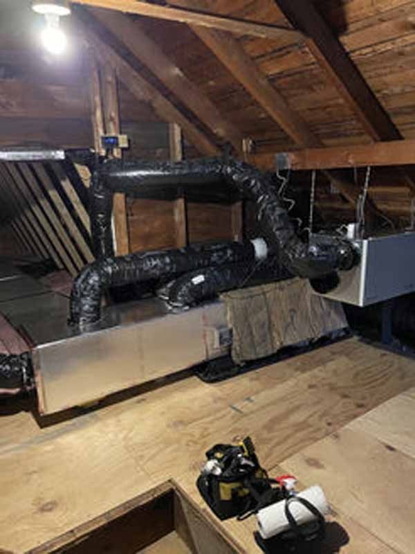 HVAC system in an attic, with insulated black ducts and a silver unit on wooden planks, with a lightbulb overhead.