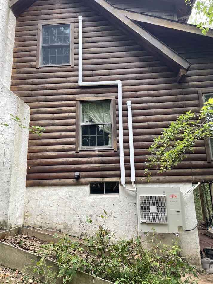Side of a log cabin with white pipes running from an air conditioning unit to above the windows.