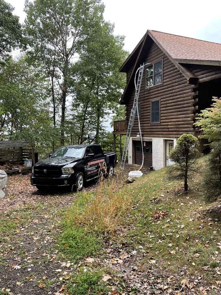 Black truck parked near a log cabin with a ladder leaning against the side, on a wooded property.