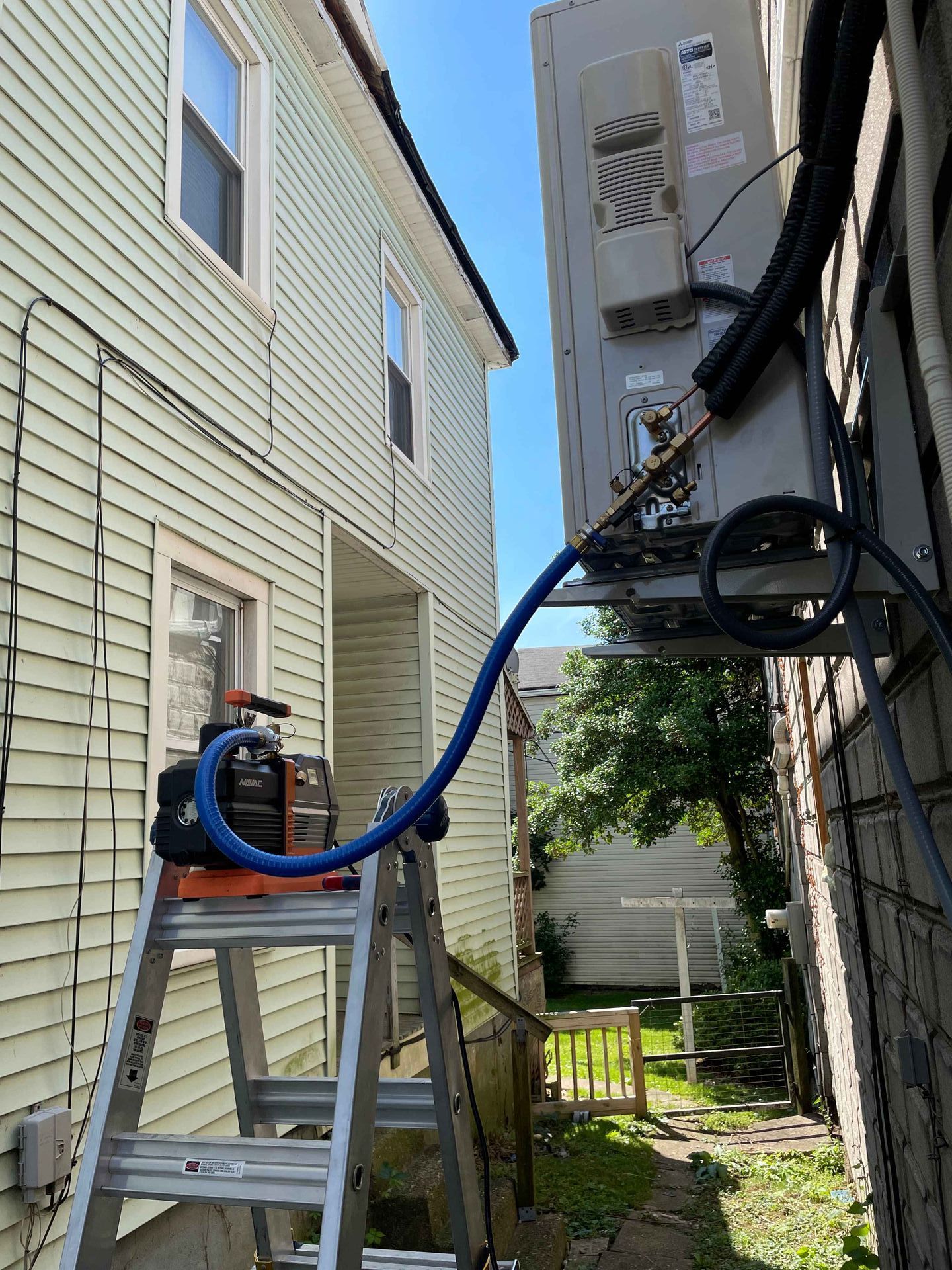 A vacuum pump on a ladder is connected to an AC unit mounted on a building's exterior.