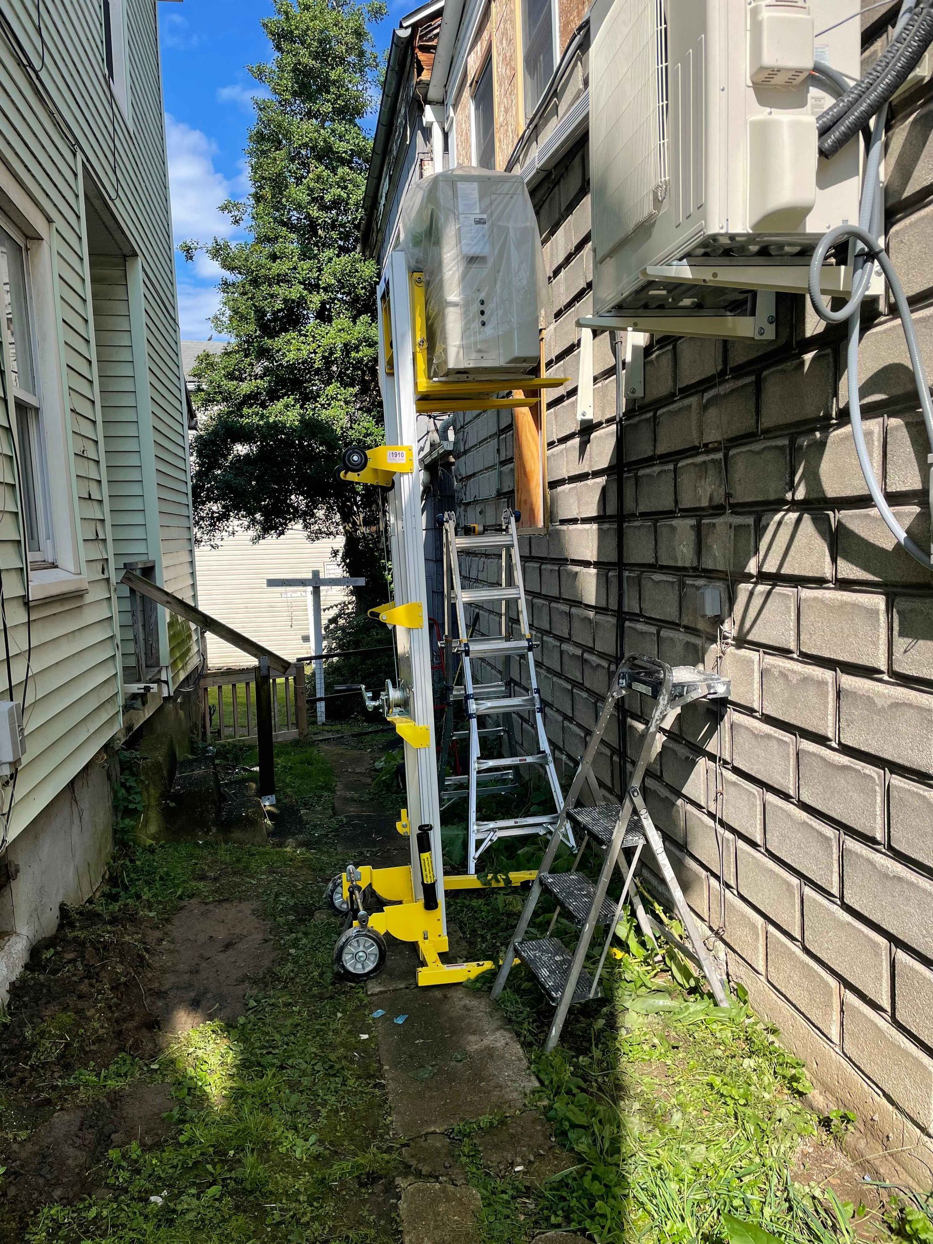Ladder propped against a brick wall next to a building, with a narrow alleyway, air conditioning units, and a grassy area.