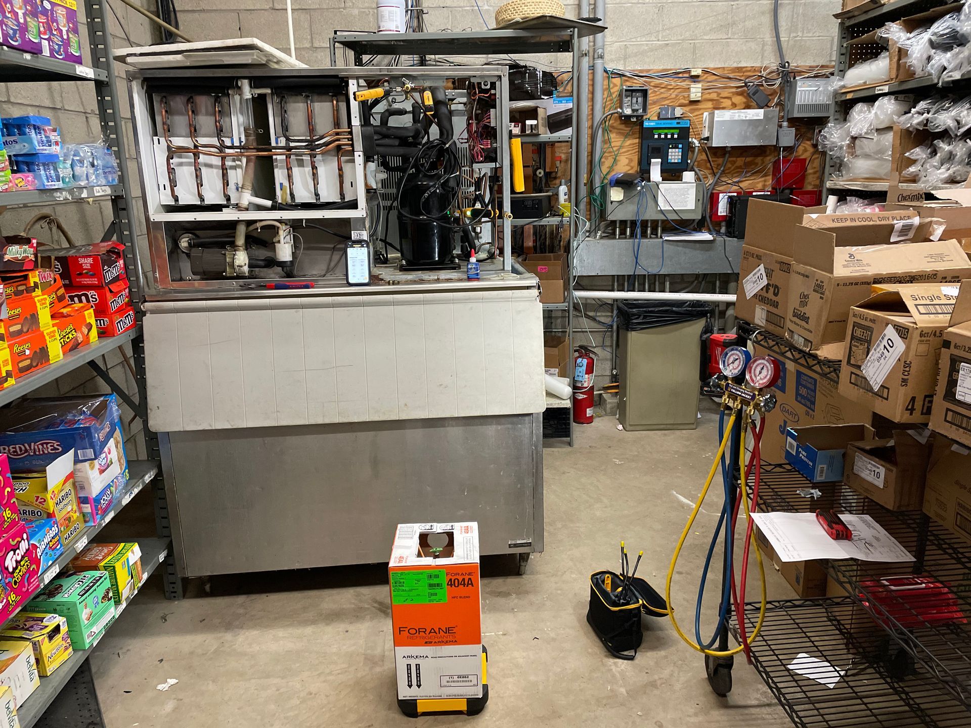 A cluttered equipment room with refrigeration units, boxes, and tools; a mechanic works.