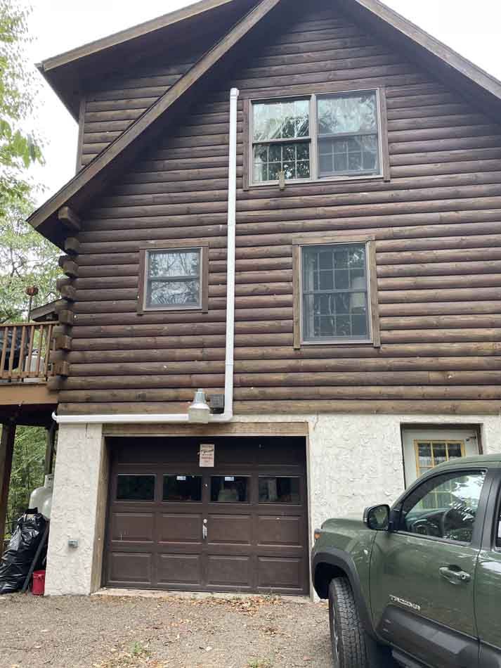 Two-story log cabin with brown garage door and white pipe. A green truck is parked nearby.