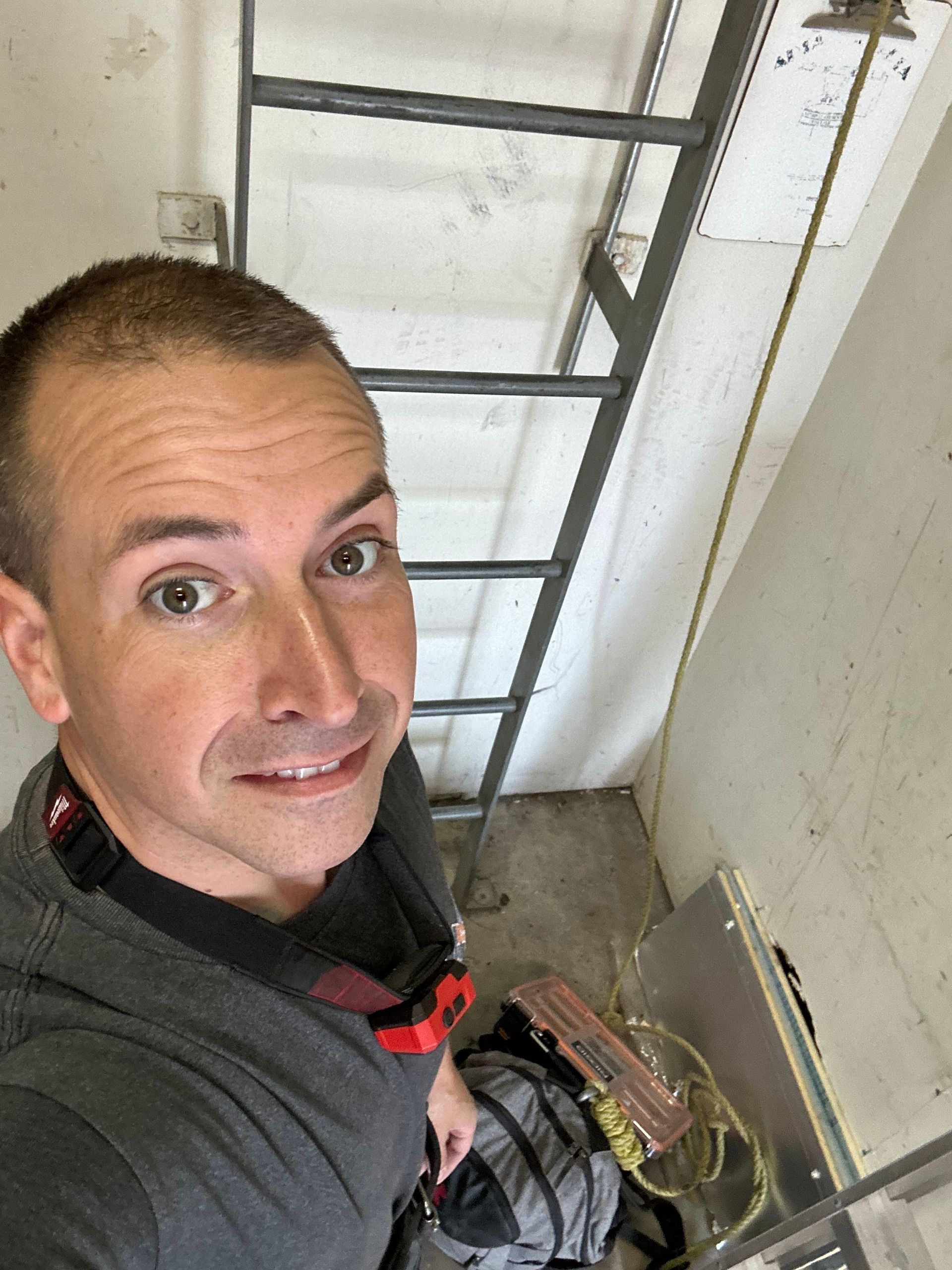Man in grey shirt smiles, taking selfie inside a structure with a ladder.